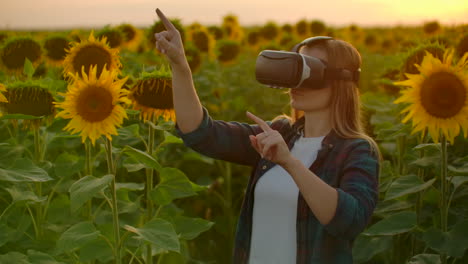 The-female-student-with-long-hair-in-plaid-shirt-and-jeans-is-working-in-VR-glasses.-She-is-engaged-in-the-working-process.-It-is-a-great-sunny-evening-in-the-sunflower-field-at-sunset.